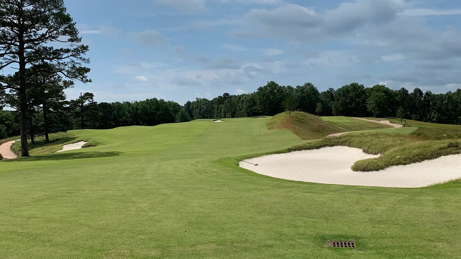 A rolling fairway climbing toward a green, flanked by large sand bunkers and tall pines under a big cloud-streaked sky.